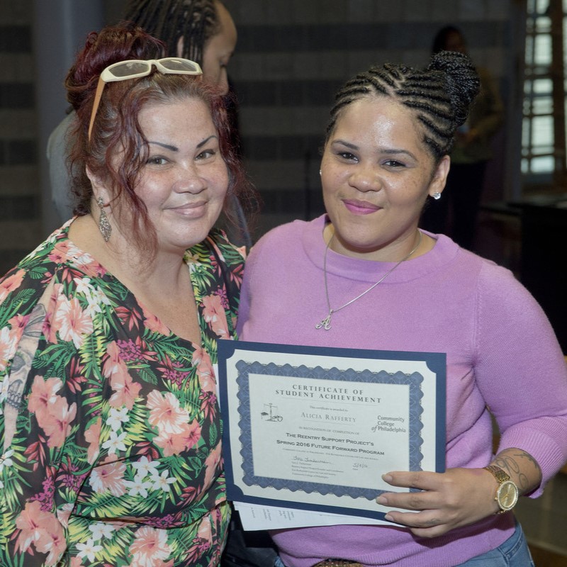 A young women holds a diploma as she stands with her mother